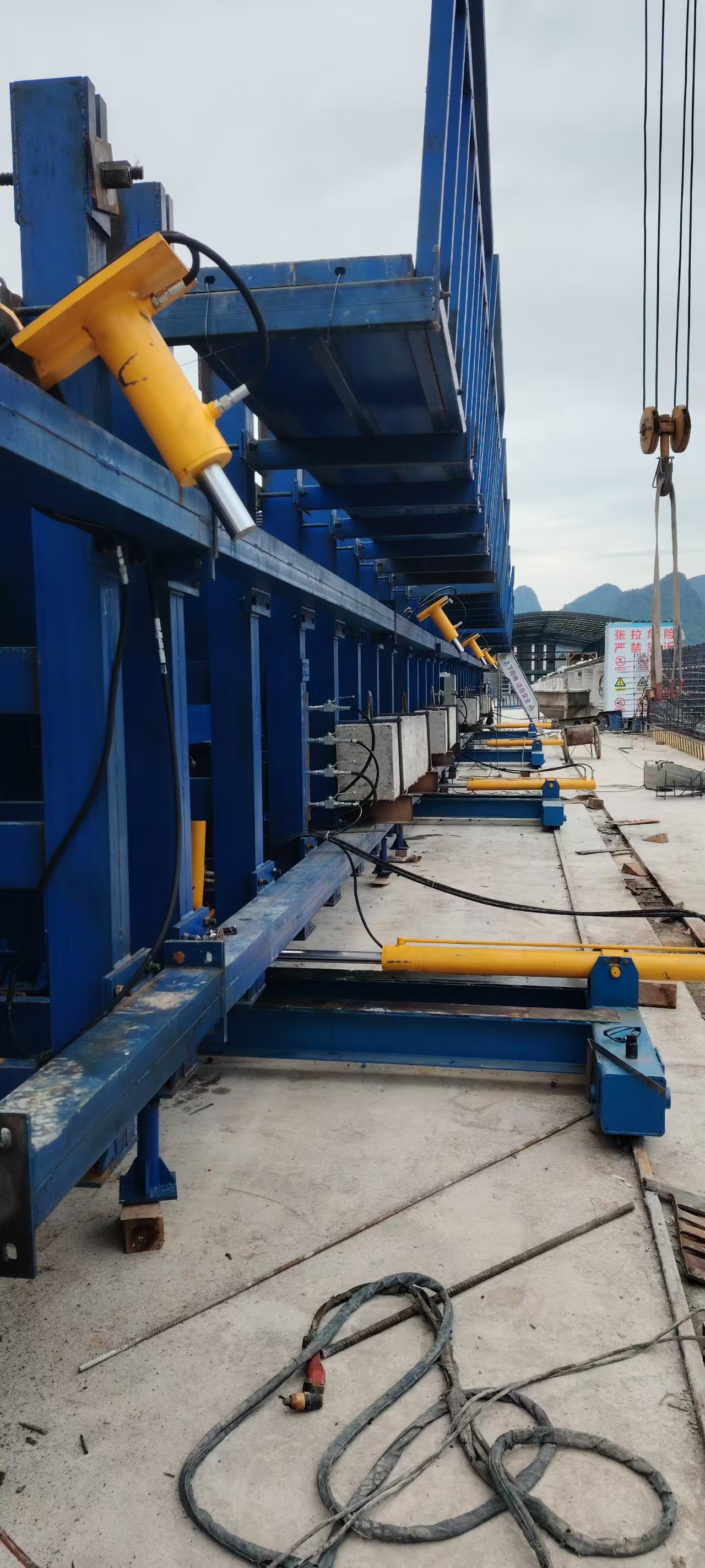 Yellow hydraulic cylinders connected to long blue steel side formwork at a precast bridge yard